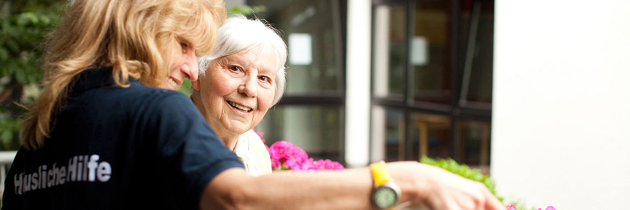 Eine lachende Seniorin mit einer DRK-Mitarbeiterin beim Blumen gießen. 